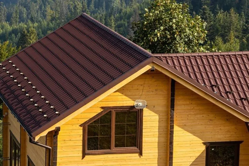 Close-up view of a wooden house with a brown metal tile roof, surrounded by a lush green forest, showcasing durability and aesthetic charm